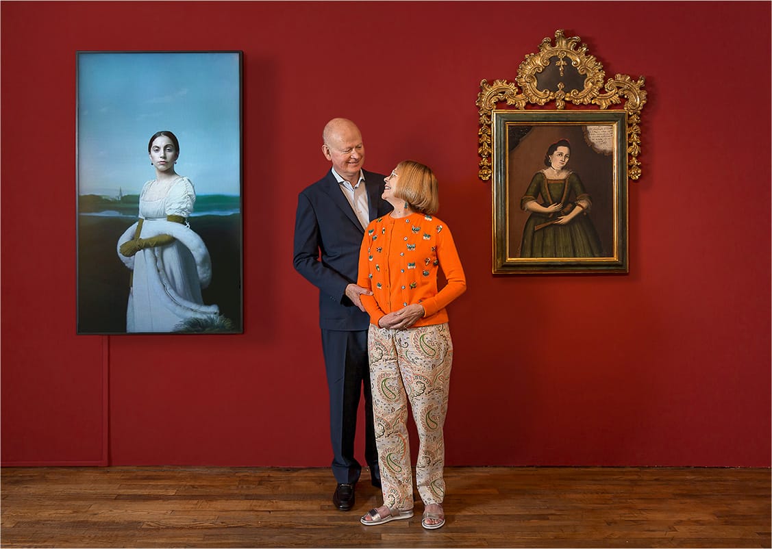 A white couple in nice clothing smiling at each other in front of a red wall with a historic painting and digital artwork.
