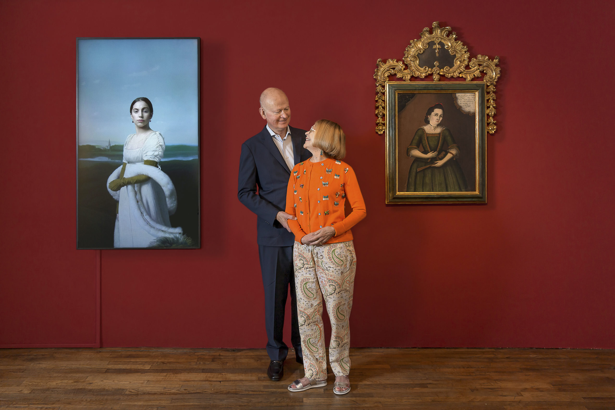 Carl and Marilynn Thoma standing in front of a red wall with two works of art.