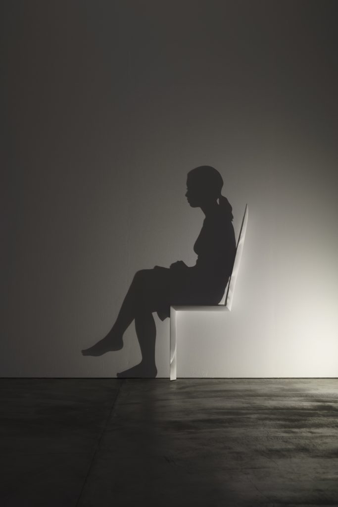 A wall sculpture comprised of cast light and its shadow depicts a woman seated in a chair.