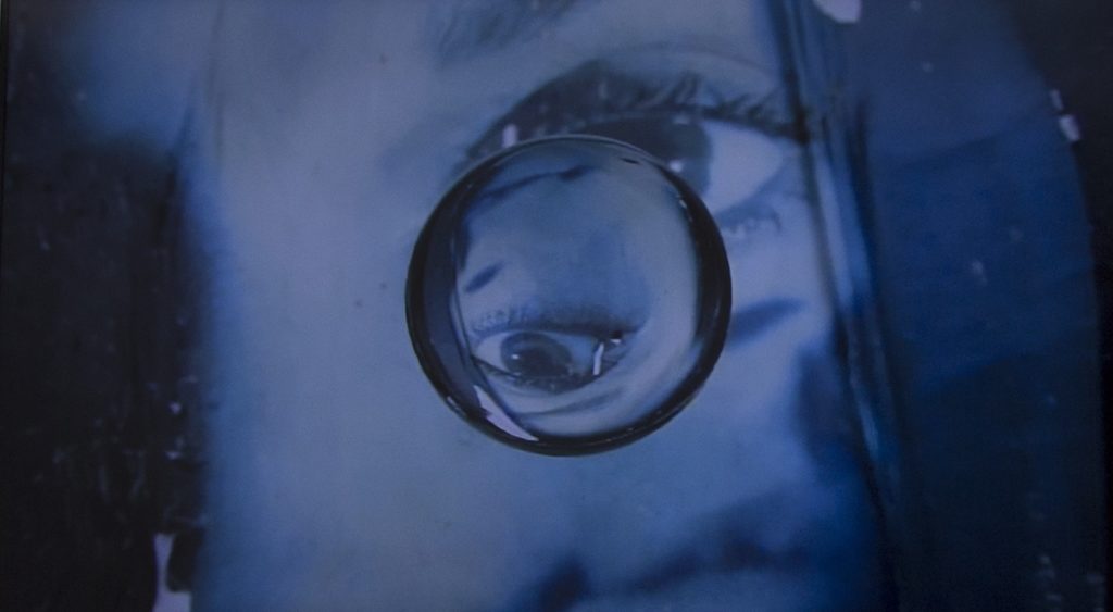 A video still of a bluish scene depicting a close up of a woman's face. In the center of the scene, a floating orb inverts the image behind.
