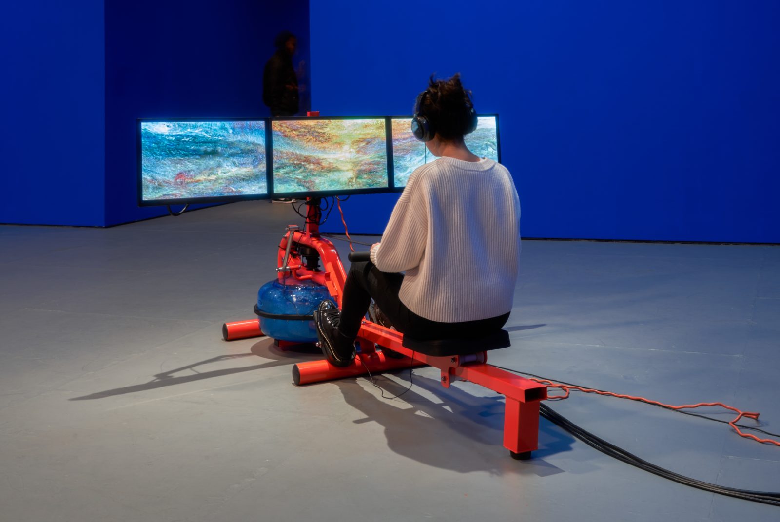 A woman in a white sweater, and wearing headphones, sitting on an orange rowing machine with three monitors depicting waves. The room around her is a vibrant blue.