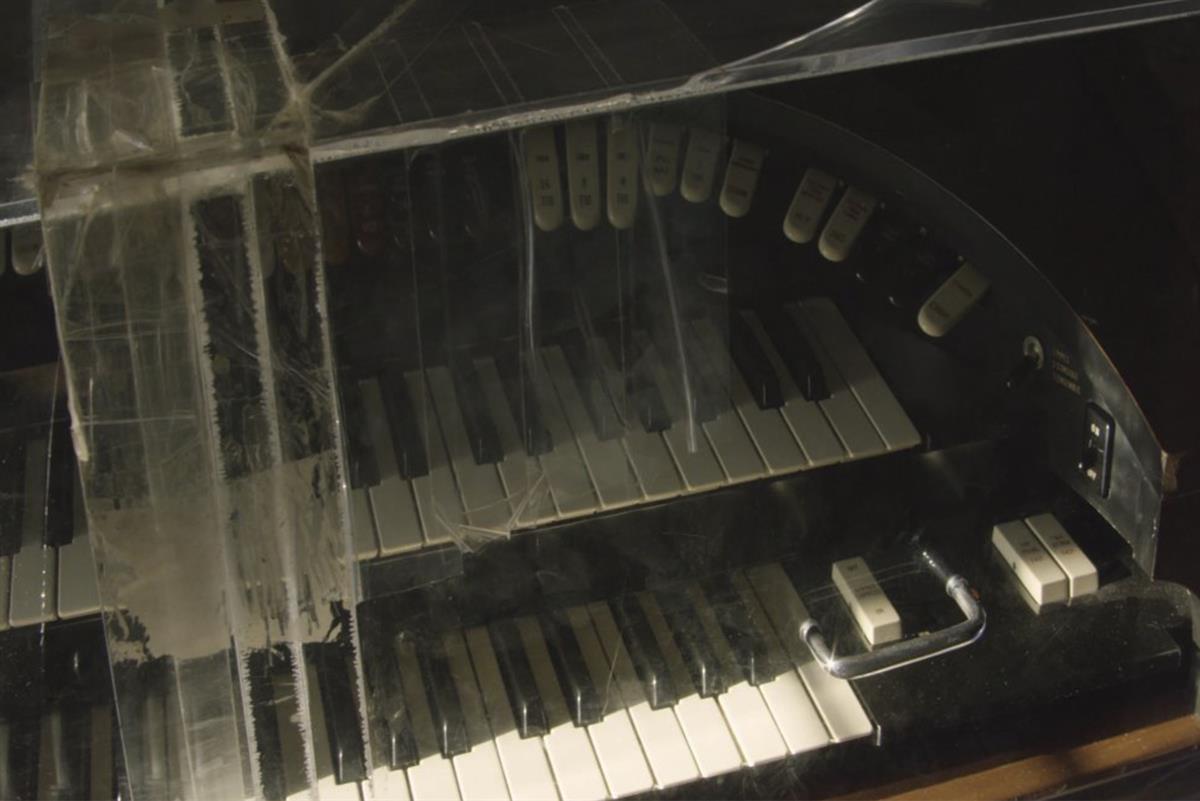 A video still depicting the right part of a piano encased in an acrylic cover with clear tape over parts of it.